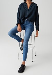 Femme portant un chemisier sombre à manches longues, un jean skinny bleu et des mocassins noirs, assise sur un tabouret en métal argenté.