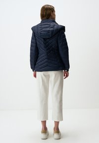 Giacca imbottita navy a trama a zigzag con cappuccio, caratterizzata da un motivo a chevron e dettagli con bottoni. Abbinata a pantaloni bianchi cropped e sneakers beige.