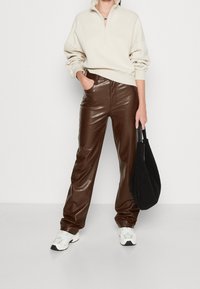 Jersey de cremallera corta color crema, pantalones marrones de piel sintética, zapatillas blancas y un bolso negro con textura. Enfoque en los contrastes de material y color.