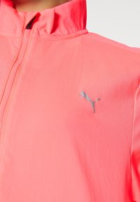 Koraleowy różowy zamek-kurtka z wysokim kołnierzem i małym odblaskowym logo Pumy po lewej stronie klatki piersiowej.