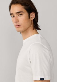 Jeune homme aux cheveux bruns mi-longs portant un t-shirt blanc Hackett London, regardant sur le côté sur un fond clair uni.