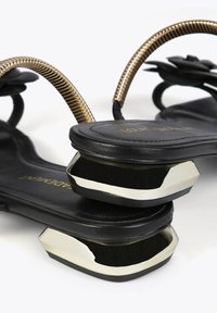 Las sandalias negras tienen un diseño elegante con una correa de tobillo metálica dorada y un acento geométrico plateado en el tacón, mostrando una textura suave.