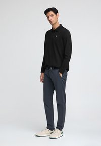 Svart långärmad polotröja, slim-fit marinblå byxor och krämfärgade sneakers med gröna accenter. Slät tygkvalitet och subtil logotypdetalj.