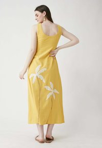 Gelbes Leinenkleid mit rundem Rückenausschnitt, das seitlich mit weißen gestickten Palmenbaum-Motiven und einer Schlitznaht versehen ist.