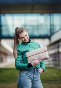 Béžová kabelka na ruku s překlopným víkem, s hladkým povrchem a jemným logem. Modelka má na sobě zelenou prošívanou bundu.