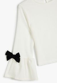 Top bianco testurizzato con maniche a campana arricciate e un nastro in tessuto nero decorativo al polso. Collo semplice con chiusura a bottone sul retro.