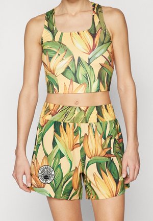 Mouwloze cropped top en shorts met geel-groene tropische bloemenprint gedragen door een staand persoon.