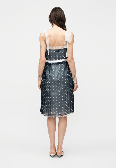 Femme portant une robe bleu marine transparente à pois blancs, bretelles nouées aux épaules, bordure en dentelle, et chaussures blanches à talons moyens, vue de dos.