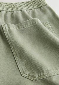 Tissu vert clair avec une ceinture élastique et une poche carrée cousue, montrant une texture et des coutures détaillées.