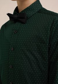 Mörkgrön knapp-up skjorta med prickigt mönster av små vita cirklar. Inkluderar en matchande svart fluga och en bröstficka.