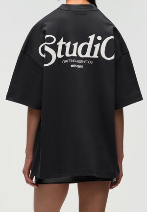 Persoon die een oversized zwart T-shirt draagt met witte tekst "Studio Crafting Aesthetics WRSTBHVR" op de rug tegen een effen achtergrond.