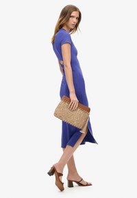 Dopasowana sukienka w królewskim niebieskim kolorze z krótkimi rękawami i wycięciem. Modelka trzyma plecioną kopertówkę z brązowym skórzanym wykończeniem i frędzlami. Brązowe sandały na obcasie.