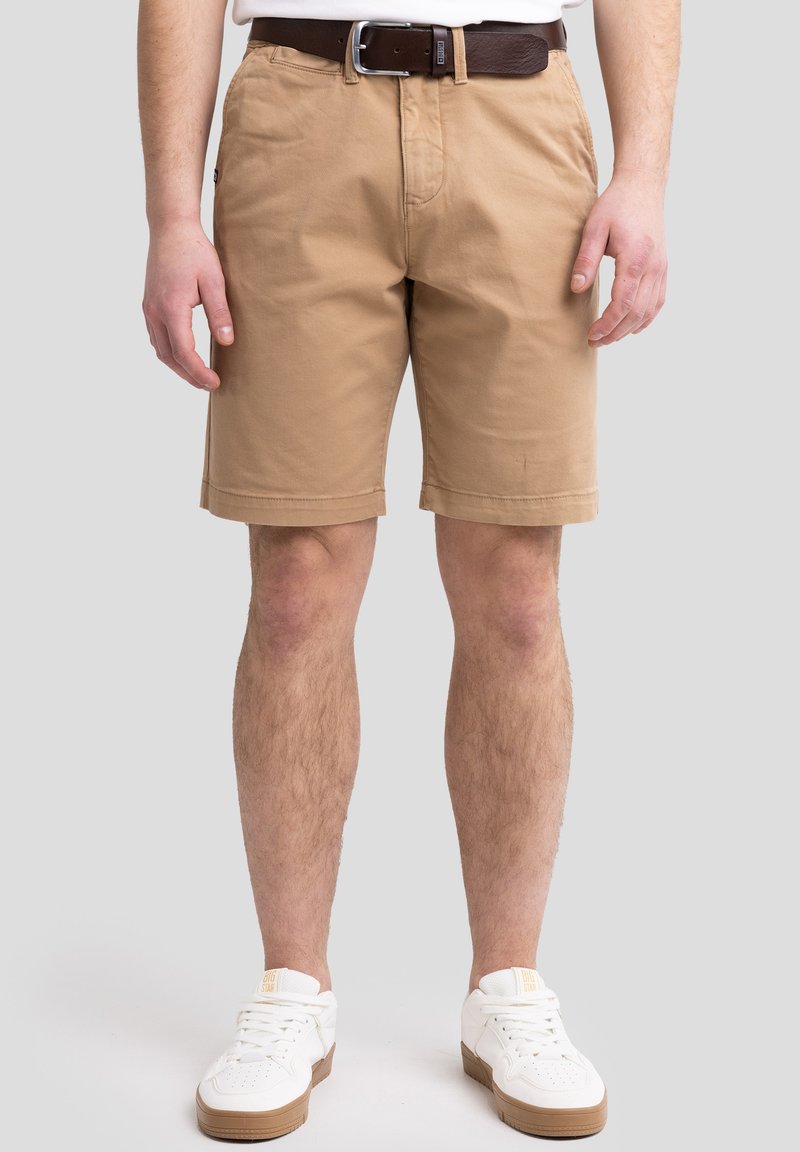 Beige Baumwollshorts mit umgeschlagenen Säumen, seitlichen Taschen und einem Gürtel. Tragen Sie dazu weiße Sneakers. Einfaches Design mit glatter Textur.