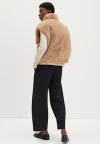 Beige Weste aus Kunstfell ohne Ärmel mit hohem Kragen, über einem hellen Pullover getragen, kombiniert mit lockeren schwarzen Hosen und schwarzen Schuhen.