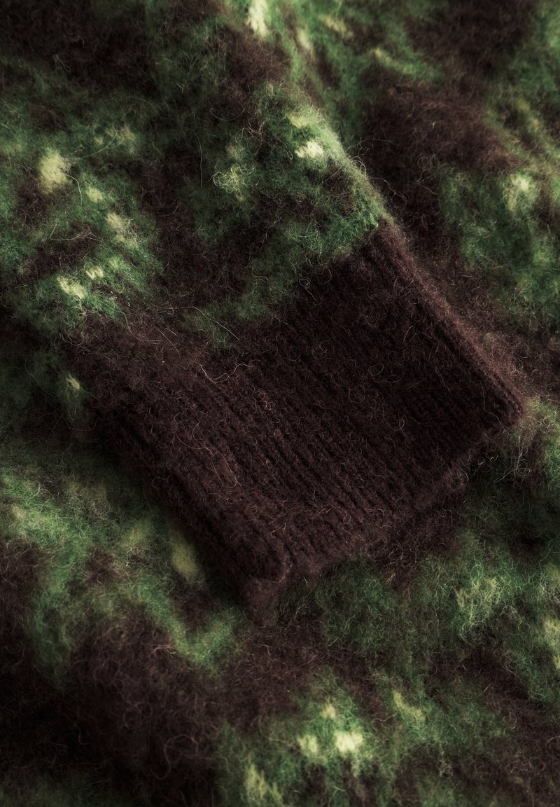 Un pull en tricot arborant un motif de camouflage vert et marron avec un poignet texturé marron foncé, fabriqué à partir d'un matériau en laine moelleuse.