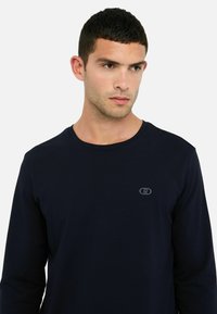 T-shirt a maniche lunghe blu navy in morbido cotone con scollatura a girocollo e un sottile dettaglio del logo sul petto. Tessuto liscio, vestibilità regolare.
