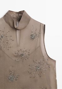 Mouwloze doorzichtige taupekleurige blouse met hoge kraag en zilveren kralen zonnestraalversieringen verspreid aan de voorkant.