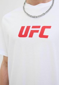 Balta medvilnės marškinėliai su ryškiai raudonu "UFC" logotipu per krūtinę. Dizainas paprastas, su trumpomis rankovėmis ir apvaliu kaklu.