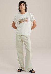 White cotton t-shirt featuring "MIAMI" in colorful printed letters. Paired with light beige wide-leg pants and black sandals with a thick strap.