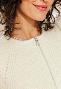Pull en tricot crème avec col montant, fermeture éclair argentée et texture douce ; possède un motif vertical subtil et un design léger.