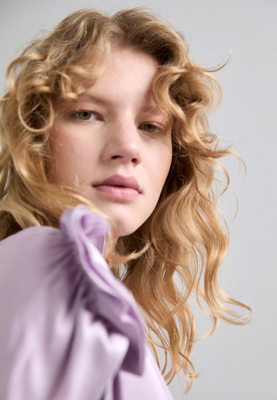 Lila bluza z nabrano detajema na rokavih, mehka tkanina in sproščen kroj. Valoviti, do ramen dolgi blond lasje na nevtralni ozadju.