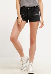 Persona vistiendo pantalones cortos de mezclilla negra con bolsillos con cremallera en el frente, camiseta gris metida por dentro, y zapatillas altas blancas, de pie con una pierna ligeramente levantada.