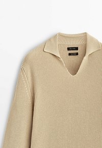 Beżowy sweter z dzianiny z dekoltem w serek i kołnierzem. Posiada teksturowany wzór i lekko luźny krój. Wewnątrz przy dekolcie widoczne metki marki.