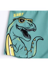 Turquoise zwembroek met een geprinte T. rex in een zonnebril, die een gele surfplank vasthoudt. Het ontwerp bevat zwarte omtrekken en accenten.