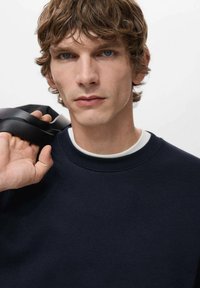 Navyblauwe trui met een ronde hals en geribbelde manchetten, gedragen over een wit overhemd. Het model houdt een zwarte band over één schouder vast.