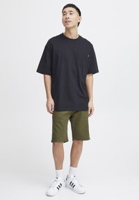 Schwarzes Baumwoll-T-Shirt mit Tasche, kombiniert mit olivgrünen Shorts. Dazu weiße Sneaker mit schwarzen Streifen und weiße Crew-Socken.