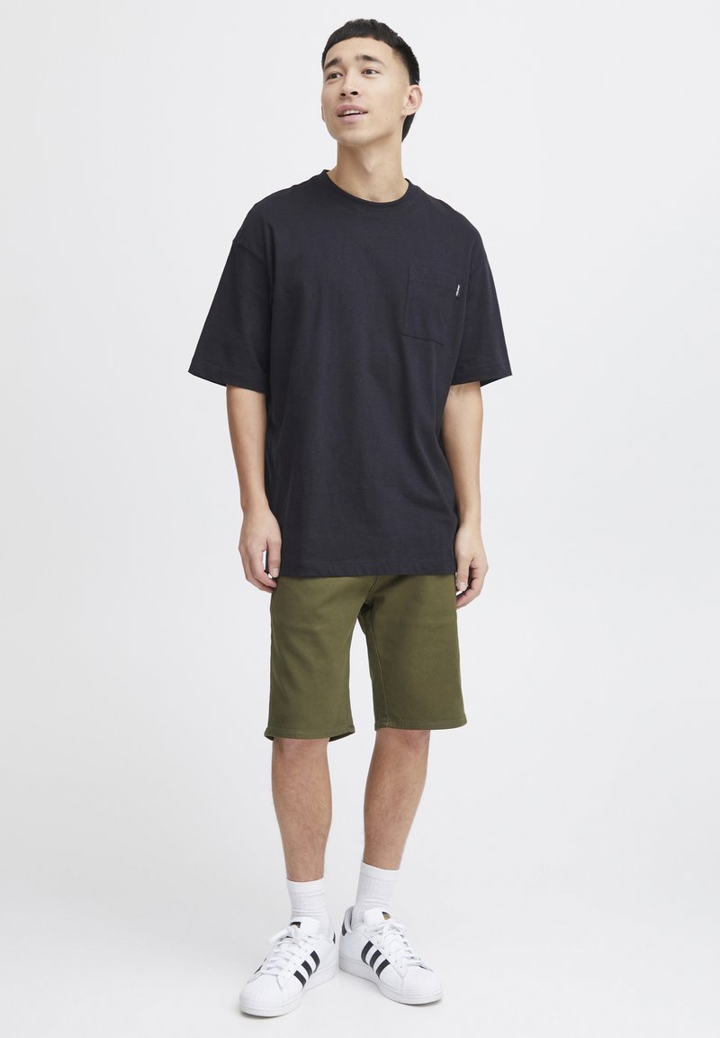 Schwarzes Baumwoll-T-Shirt mit Tasche, kombiniert mit olivgrünen Shorts. Dazu weiße Sneaker mit schwarzen Streifen und weiße Crew-Socken.