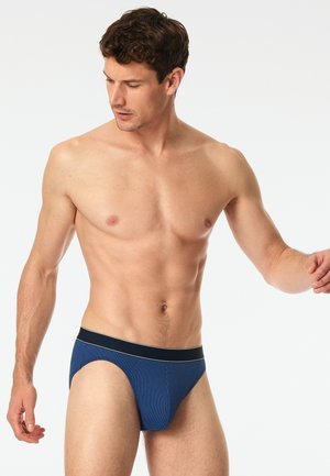 Blaue gerippte Slips mit einem schwarzen Bund, der ein dezentes Logo und weiße Akzentlinien aufweist. Das gezeigte Modell steht mit erhobener Arm.