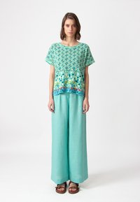 Korte mouwen shirt in turquoise met geometrische patronen, gecombineerd met losse, lichtblauwe linnen broek. Model draagt bruine sandalen.