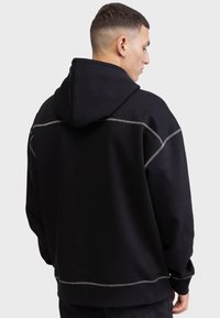 Juodas hoodie, pasiūtas iš tekstūruotos medžiagos, su dideliu gobtuvu ir kontrastingomis siūlėmis, išryškinančiomis šonus ir apatinę dalį. Rodyma atgal.