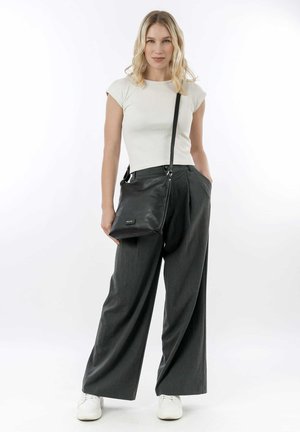 Zwarte leren handtas met verstelbare schouderriem, een gladde textuur en subtiele branding. Model draagt een strakke witte top en grijze wijde broeken.