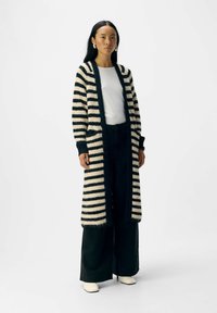 Langer gestreifter Cardigan in Creme und Schwarz, gestrickte Textur, mit offener Front und gerippten Manschetten, getragen über einem weißen Oberteil und dunklen Hosen.