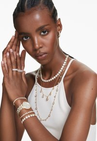 Goldfarbene Schmuckstücke mit mehreren Halsketten, Armbändern und Ringen, die mit klaren Edelsteinen verziert sind, präsentiert von einem Model in einem weißen Oberteil.