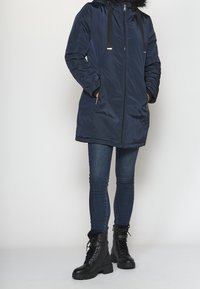 Parka acolchada azul marino con ribete de piel sintética en la capucha, cierre de cremallera y bolsillos laterales, combinada con vaqueros ajustados azul oscuro y botas negras con cordones.