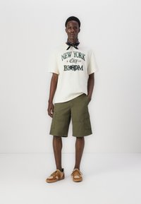 Lys beige bomulds t-shirt med grønnt tryk, der står "NEW YORK a City in BLOOM." Olivengrønne cargo shorts, brune og tan sneakers, sort bandana.