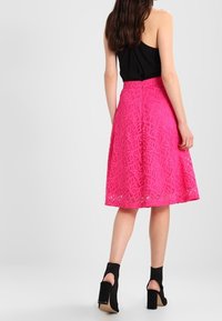 Nainen, jolla on yllään hihaton musta paita ja polven pituinen kirkkaanpinkki kukkakirjailtu pitsihame, yhdistettynä mustiin korkokenkiin, joissa on avoin takakappale.