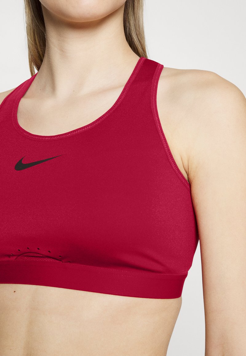 Reggiseno sportivo rosso realizzato in materiale liscio ed elastico. Presenta un logo Nike nero e un colletto arrotondato con fori di ventilazione lungo il fondo.