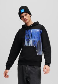 Felpa nera con stampa grafica blu che rappresenta un paesaggio urbano, con lacci, tasca frontale e abbinata a un berretto lavorato a maglia nero.