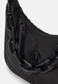 Sac en tissu matelassé noir avec une large bandoulière chaîne noire, une fermeture éclair visible et une tirette embossée "Steve Madden".