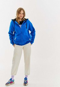 Niebieska bluza z zamkiem błyskawicznym, z czarnym wyłożonym kapturem, bocznymi kieszeniami i sznurkami; noszona na ciemnej bluzce, w zestawieniu z kremowymi spodniami i kolorowymi sneakersami.