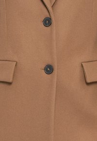 Manteau en laine couleur camel avec deux boutons noirs et deux poches à rabat à l'avant, montrant un gros plan de la taille.