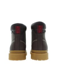 Un paio di stivali in pelle marrone con suole spesse color tan, cuciture bianche e linguette rosse con l'etichetta "U.S. Polo Assn." viste da dietro.