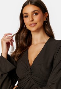 Dunkel gestreiftes Wickeloberteil mit tiefem V-Ausschnitt, langen Schlagärmeln und geknotetem Frontdetails. Das Model hat sanfte Wellenfrisur und goldene Creolen.