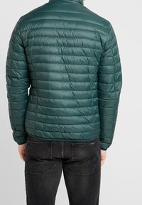 Grøn quiltet pufferjakke med høj krave, med horisontal syning og en glat, blank tekstur. Sammen med mørke jeans.
