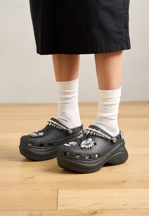 Ben med hvite sokker og svarte platåklosser dekorert med rhinesteiner og perlelignende perler, som står på et tregulv.