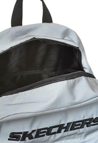 Mochila plateada con un interior negro. Cuenta con un bolsillo con cremallera, correas negras resistentes y el logo en relieve "SKECHERS" en la parte frontal.